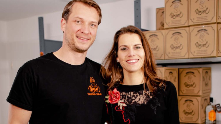 Ein Mann im schwarzen T-Shirt mit orangefarbener Aufschrift und eine Person in schwarzer Kleidung stehen vor gestapelten Kartons.