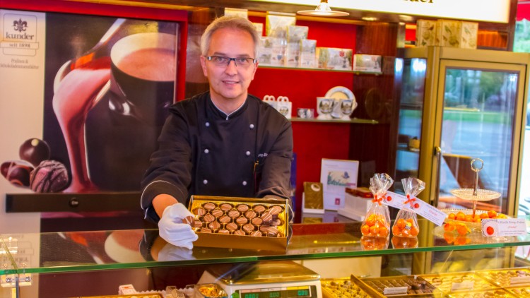 Person in schwarzer Uniform präsentiert eine Schachtel Pralinen vor einer Glasvitrine mit Schokoladen und Pralinen.