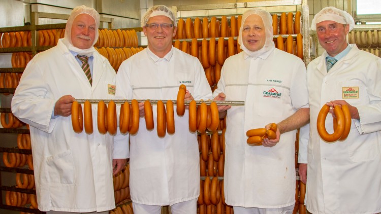 Vier Personen in weißer Arbeitskleidung halten Würstchen auf Metallstangen vor Regalen mit weiteren Würsten.