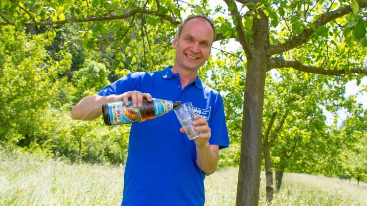 Person in blauem Shirt gießt aus einer Flasche mit der Aufschrift „Hoi!“ ein Getränk in ein Glas, im Hintergrund Bäume.