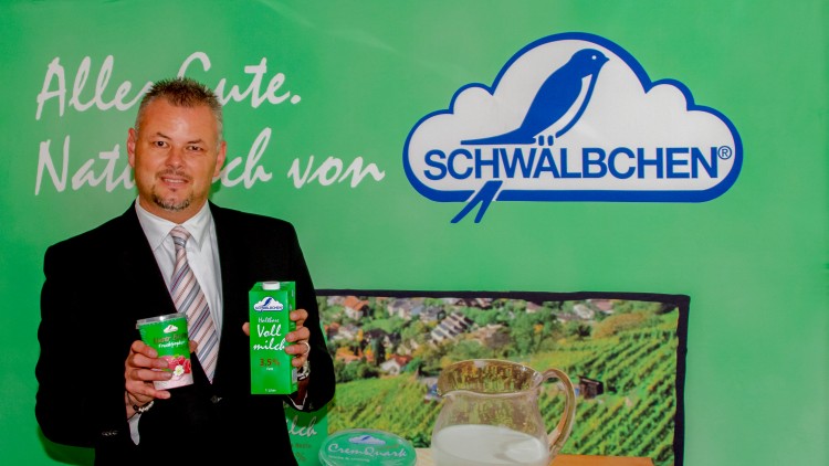 Person im Anzug hält Schwalbchen-Produkte vor grünem Hintergrund mit Logo, Milchkanne und weiteren Produkten.