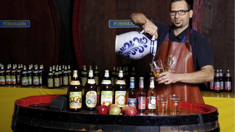 Ein Mensch schenkt Apfelwein aus einem großen Krug in ein Glas, umgeben von Flaschen, Äpfeln und Fässern mit dem Schriftzug \