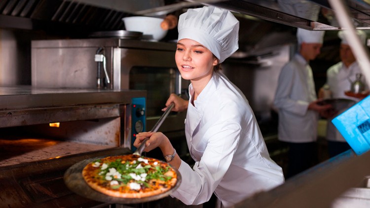 Eine Person in weißer Kochkleidung holt eine Pizza mit grünen Blättern und weißem Belag aus einem heißen Ofen.