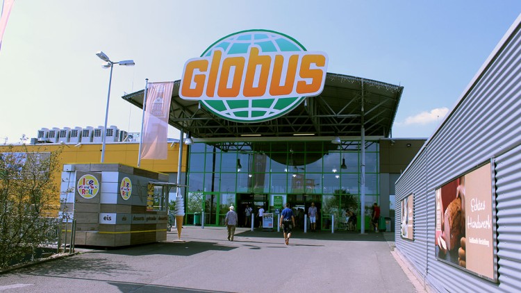 Eingangsbereich eines GLOBUS Marktes mit großem GLOBUS Logo, Eisstand links und Werbung mit der Aufschrift „Echtes Handwerk“ rechts.
