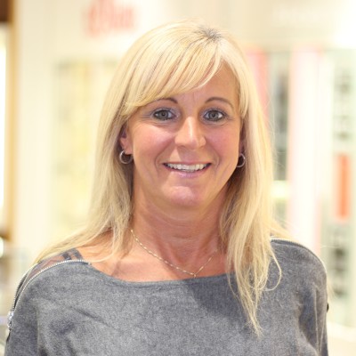 Person mit langen blonden Haaren trägt silberne Ohrringe und eine graue Oberbekleidung, Hintergrund mit heller Beleuchtung.