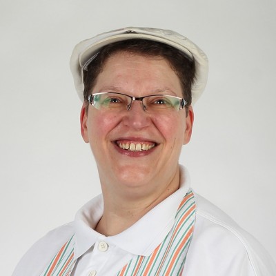 Person trägt eine weiße Schiebermütze, ein weißes Hemd und eine gestreifte Schürze in Orange, Grün und Weiß.