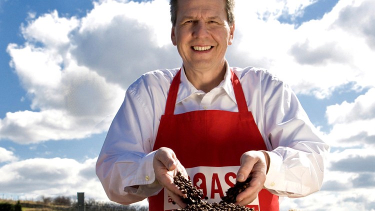 Person mit roter Schürze hält Kaffeebohnen aus einem vollen Jutesack vor einer sonnigen Landschaft mit Wolken am Himmel.