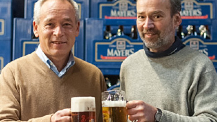 Zwei Personen halten Biergläser vor stapelweise blauen Getränkekisten mit dem Schriftzug „Mayers“.
