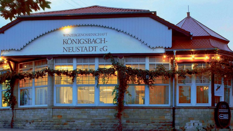 Gebäude mit rotem Dach, Glockenformverzierung, Schriftzug „Winzergenossenschaft Königsbach-Neustadt eG“, beleuchtete Fenster.