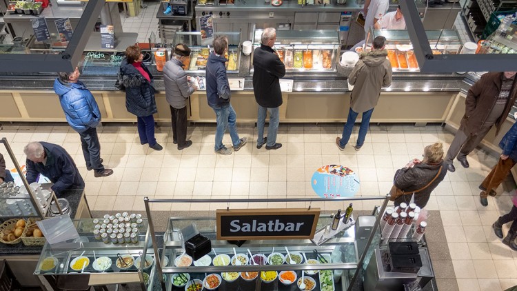 Menschen stehen in einer Selbstbedienungstheke-Schlange, daneben Getränkebereich mit Gläsern und Salatbar mit verschiedenen Speisen.