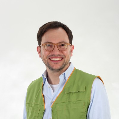 Person trägt eine grüne Weste mit orangen Nähten über einem hellblauen Hemd und einer Jeans vor hellem Hintergrund.