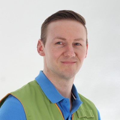 Person trägt eine grüne Arbeitsweste mit oranger Umrandung und ein blaues Poloshirt vor neutralem Hintergrund.