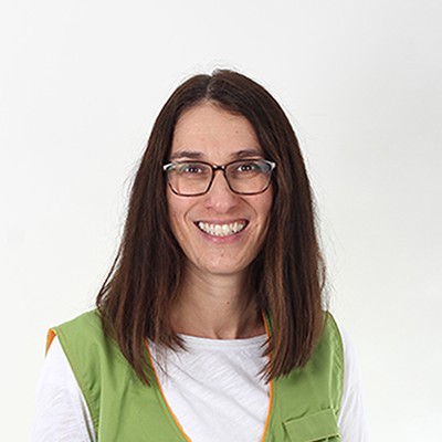 Person in weißem Langarmshirt, grauer Hose und grünem GLOBUS-Mitarbeiterwesten mit orangen Details vor hellem Hintergrund.