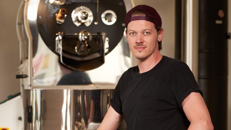 Person in schwarzem T-Shirt vor einer silbernen Kaffeeröstmaschine, umgeben von Kaffeesäcken mit Beschriftung und Metallschaufel.
