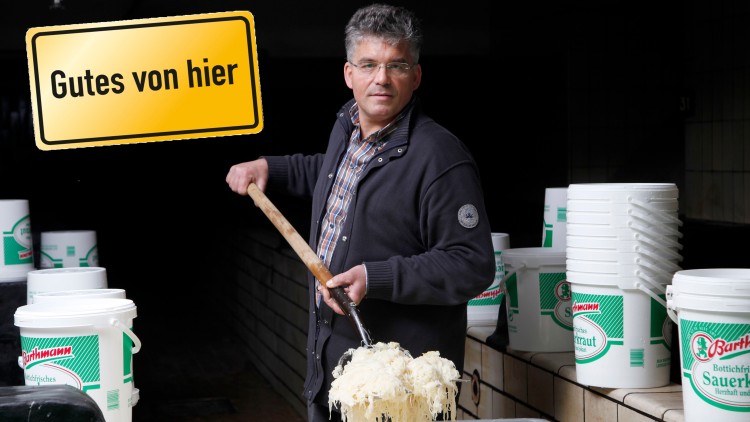 Person rührt mit einem Holzstab in einer Wanne mit Sauerkraut, daneben stehen Eimer mit der Aufschrift „Bartmann Bottichfrisches Sauerkraut“.