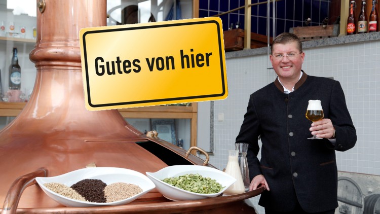 Kupferner Braukessel mit Schalen voller Getreide und Hopfen, daneben ein Mann mit Bierglas, Schildtext „Gutes von hier“.