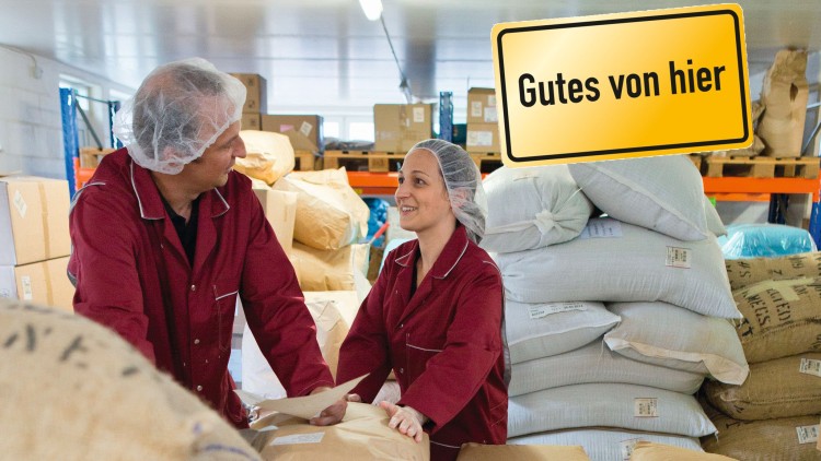 Zwei Personen in roten Kitteln stehen vor Säcken und Kartons, im Hintergrund ein gelbes Schild mit der Aufschrift Gutes von hier.