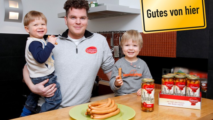 Ein Teller mit Würstchen steht auf einem Holztisch, daneben Gläser mit Frankfurter Würstchen und ein Schild mit „Gutes von hier“.