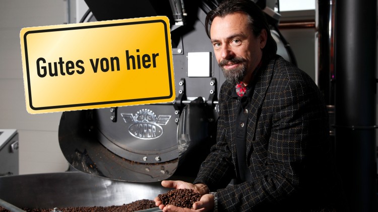 Eine Person hält geröstete Kaffeebohnen in den Händen, im Hintergrund eine Kaffeeröstmaschine und ein Schild mit „Gutes von hier“.