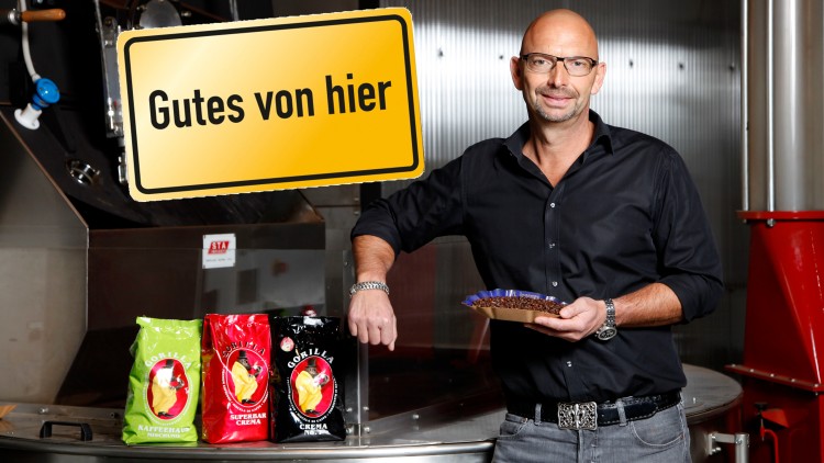 Eine Person steht vor einer Kaffeeröstmaschine mit drei Kaffeepackungen der Marke Gorilla und einem gelben Schild mit „Gutes von hier“.