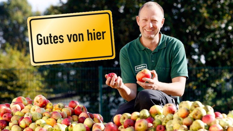 Person in grüner Kleidung hält zwei Äpfel vor einem großen Haufen Äpfel, daneben Schild mit Text „Gutes von hier“.