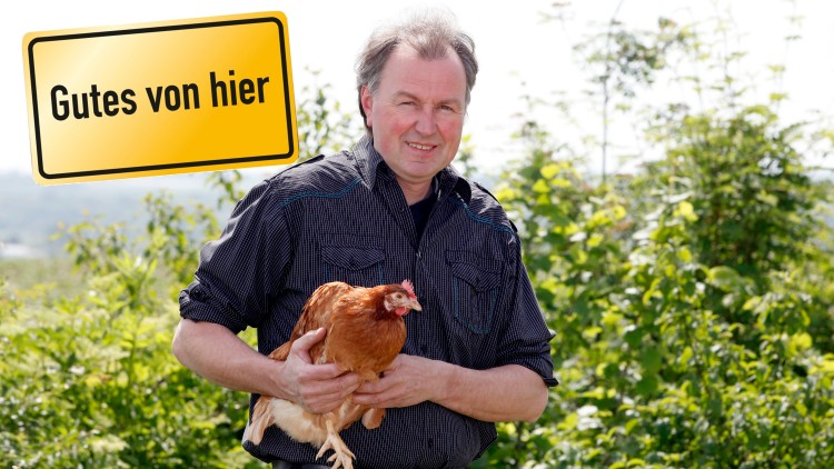 Person hält ein braunes Huhn vor grünem Hintergrund, links oben ist ein gelbes Schild mit der Aufschrift Gutes von hier.