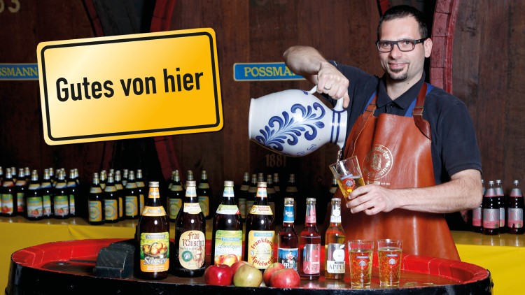 Mann trägt Lederschürze, gießt Getränk aus blau-weißem Krug in Glas auf Fass mit Flaschen, Äpfeln und Text \