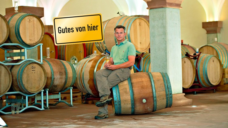Mann sitzt auf einem Holzfass in einem Raum mit mehreren Weinfässern, davor ein gelbes Schild mit der Aufschrift Gutes von hier.