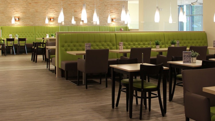 Helles Restaurant mit grünen Polsterbänken, Holzstühlen, Tischen sowie Pflanzen und Karten als Tischdekoration.