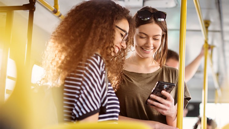 Person hält ein Smartphone in einer Straßenbahn, gelbe Haltestangen und unscharfer Hintergrund mit einer Tür sind sichtbar.