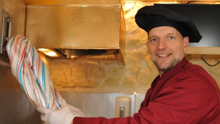 Person in roter Kleidung und schwarzem Hut hält eine große, bunte Zuckerstange in einer Küche mit goldenen und beige Elementen.