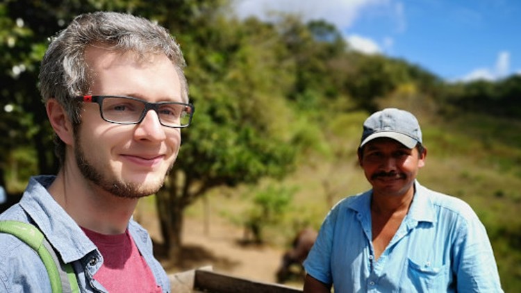 Zwei Personen stehen auf einer Wiese mit Bäumen im Hintergrund, eine trägt ein Hemd und eine Kappe, die andere hat einen Rucksack.