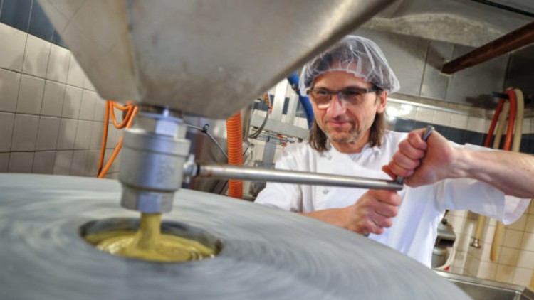 Große Metallmaschine mit drehendem Stein mahlt gelbe Masse, orangefarbene Schläuche und Kacheln im Hintergrund.