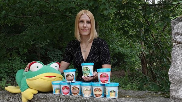 Eine Person mit einem großen Stofffrosch präsentiert auf einer Steinmauer mehrere Becher Eiscreme verschiedener Sorten.