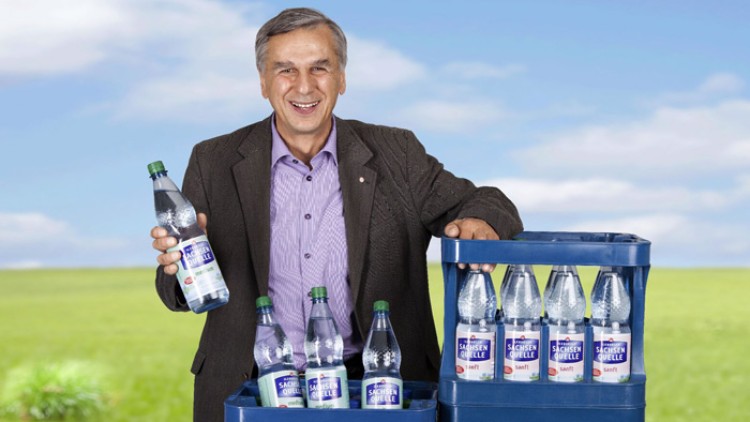Person hält eine Flasche Sprudelwasser der Marke „Schwarzwald-Sprudel“ neben zwei blauen Getränkekisten in einer Wiese.