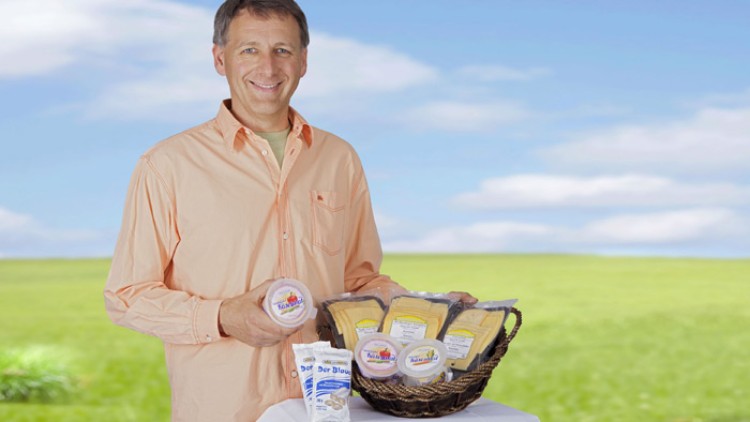 Person in beigem Hemd hält Lebensmittel, darunter Käsescheiben und ein Produkt „Quark der Blauen“, vor einer Wiese.