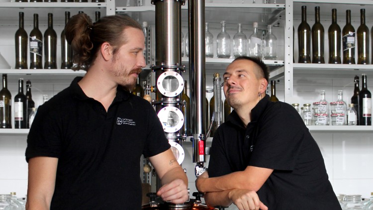 Zwei Personen in schwarzen Poloshirts mit Schriftzug vor einer Destillationsanlage und Regalen mit Flaschen und Gläsern.
