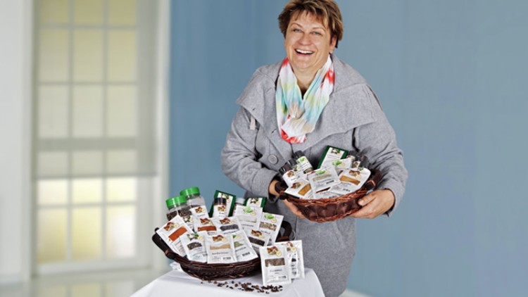 Person hält einen braunen Korb mit verpacktem Kaffee, ein weiterer gefüllter Korb steht auf einem weißen Tisch, Kaffeebohnen daneben.