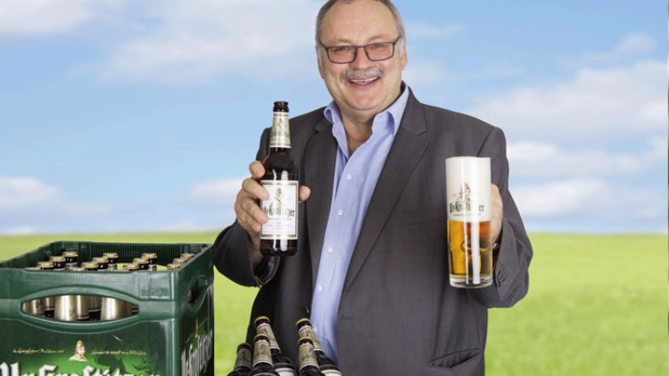 Ein Mann in Anzug hält eine Bierflasche und ein gefülltes Bierglas, daneben eine grüne Getränkekiste mit Bierflaschen.