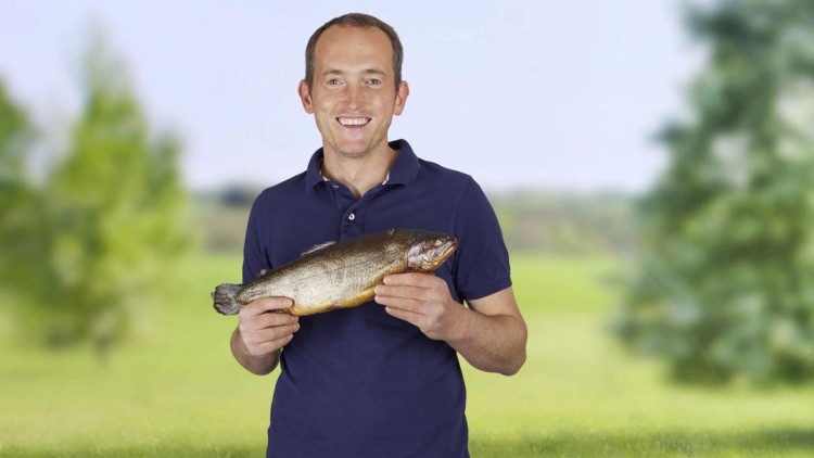 Person in blauem Poloshirt hält einen Fisch mit beiden Händen vor einer grünen Landschaft mit Bäumen.