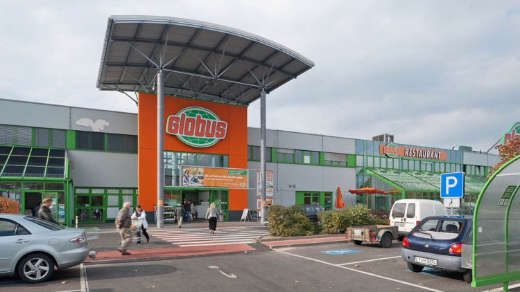 Eingangsbereich eines GLOBUS Marktes mit orangener Fassade, grünem Logo und Parkplatz mit Einkaufswagenstation.