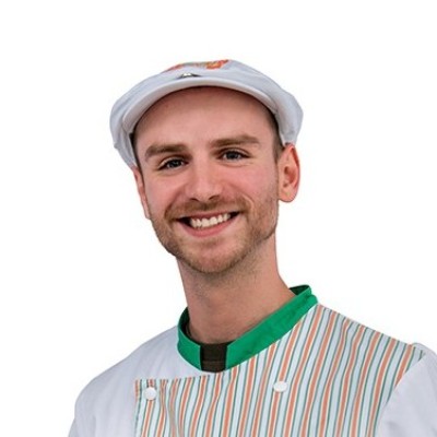 Person in weißer Arbeitskleidung mit gestreifter Schürze und grünem Kragen trägt eine weiße Mütze mit orangen Details.