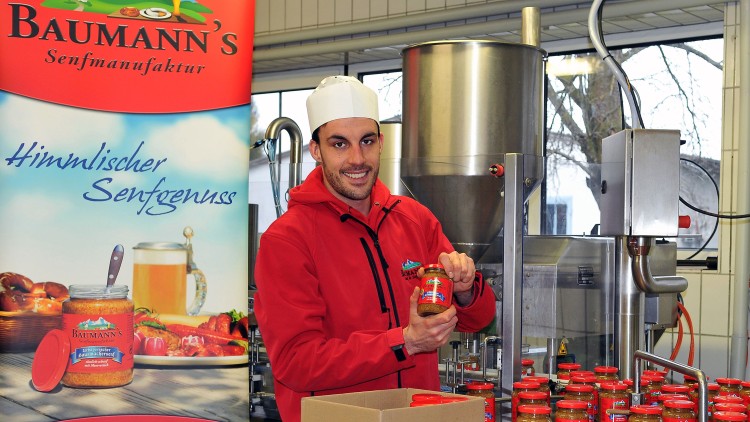 Ein Mensch in roter Jacke hält ein Glas Baumann's Senf vor Maschinen, daneben ein Banner mit „Himmlischer Senfgenuss“.