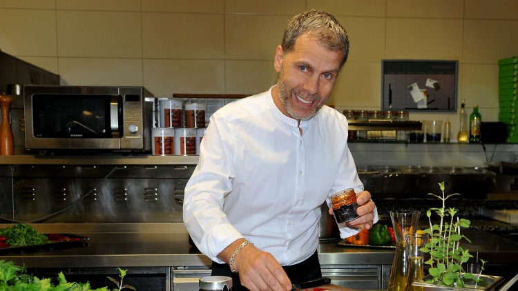 Eine Person würzt ein rohes Stück Fleisch mit einer roten Paste, umgeben von frischen Kräutern, Tomaten und Gewürzdosen.