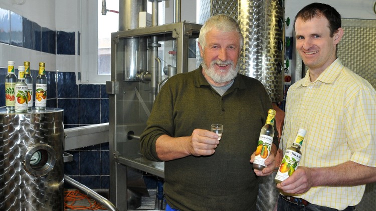 Zwei Personen stehen in einer Brennerei, halten Flaschen mit Obstbrand und ein Glas in der Hand, im Hintergrund Metalltanks.