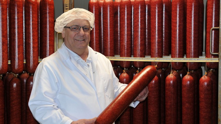 Person in weißer Arbeitskleidung hält eine große rote Wurst, im Hintergrund hängen viele Würste an Haken.