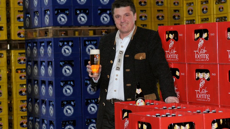 Person in Trachtenjacke hält ein Bierglas, umgeben von blauen und roten Getränkekisten mit Markenlogos.