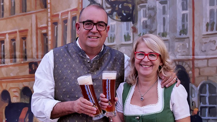 Zwei Personen in traditioneller Kleidung halten jeweils ein Glas mit dunklem Bier vor einer Kulisse mit Häuserfassaden.