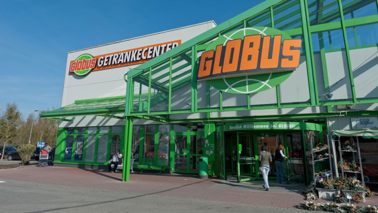 Eingangsbereich eines GLOBUS Marktes mit grünem Glasdach, großem GLOBUS-Logo, Blumenverkaufsstand und „GETRÄNKECENTER“-Schriftzug.