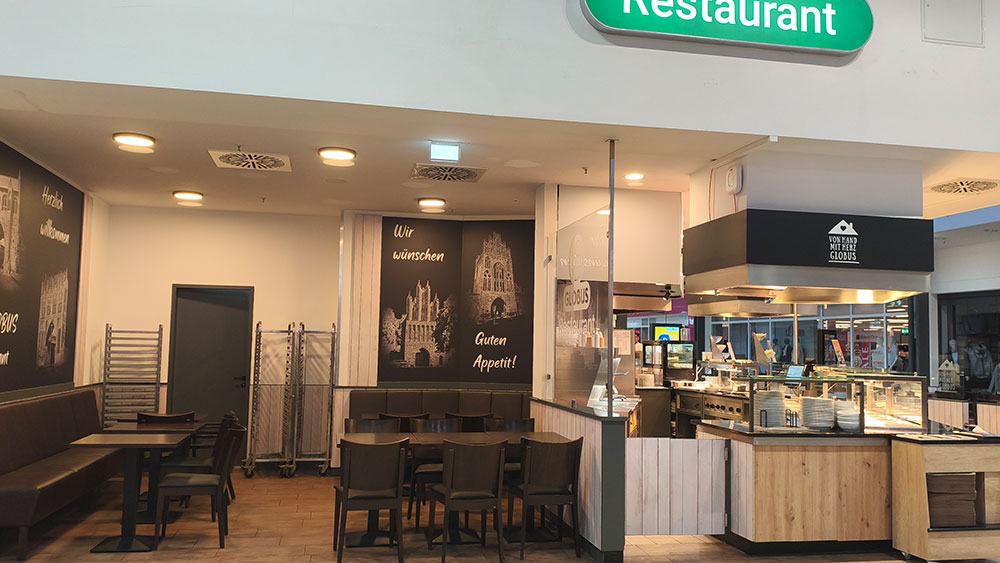 Ein Restaurantbereich mit Tischen, Stühlen, Speisetheke, Wandbildern und Beschriftungen wie „Wir wünschen Guten Appetit“.